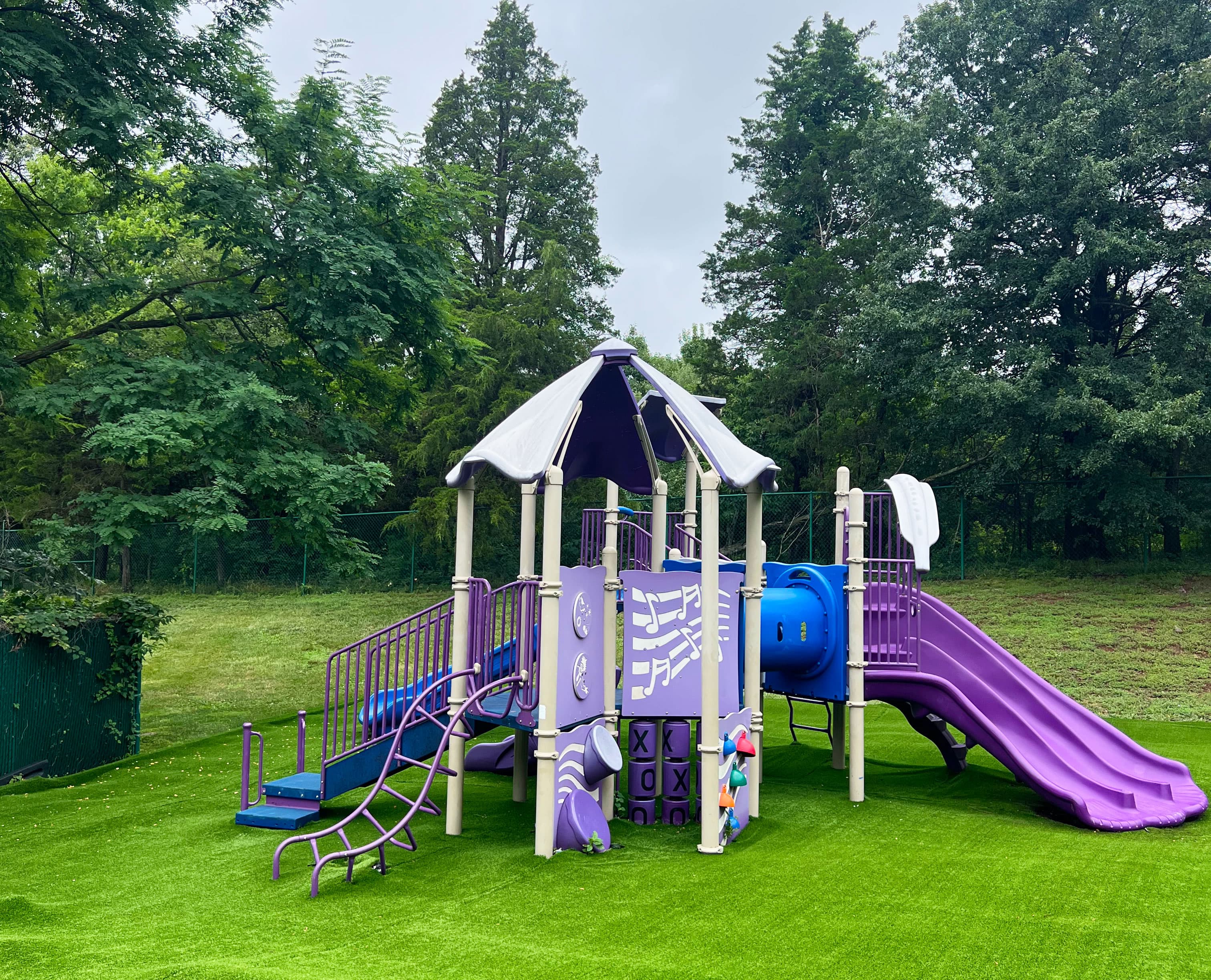 The Sterling playground with a purple slide and climbing structure on bright green turf, enclosed by a fence and trees